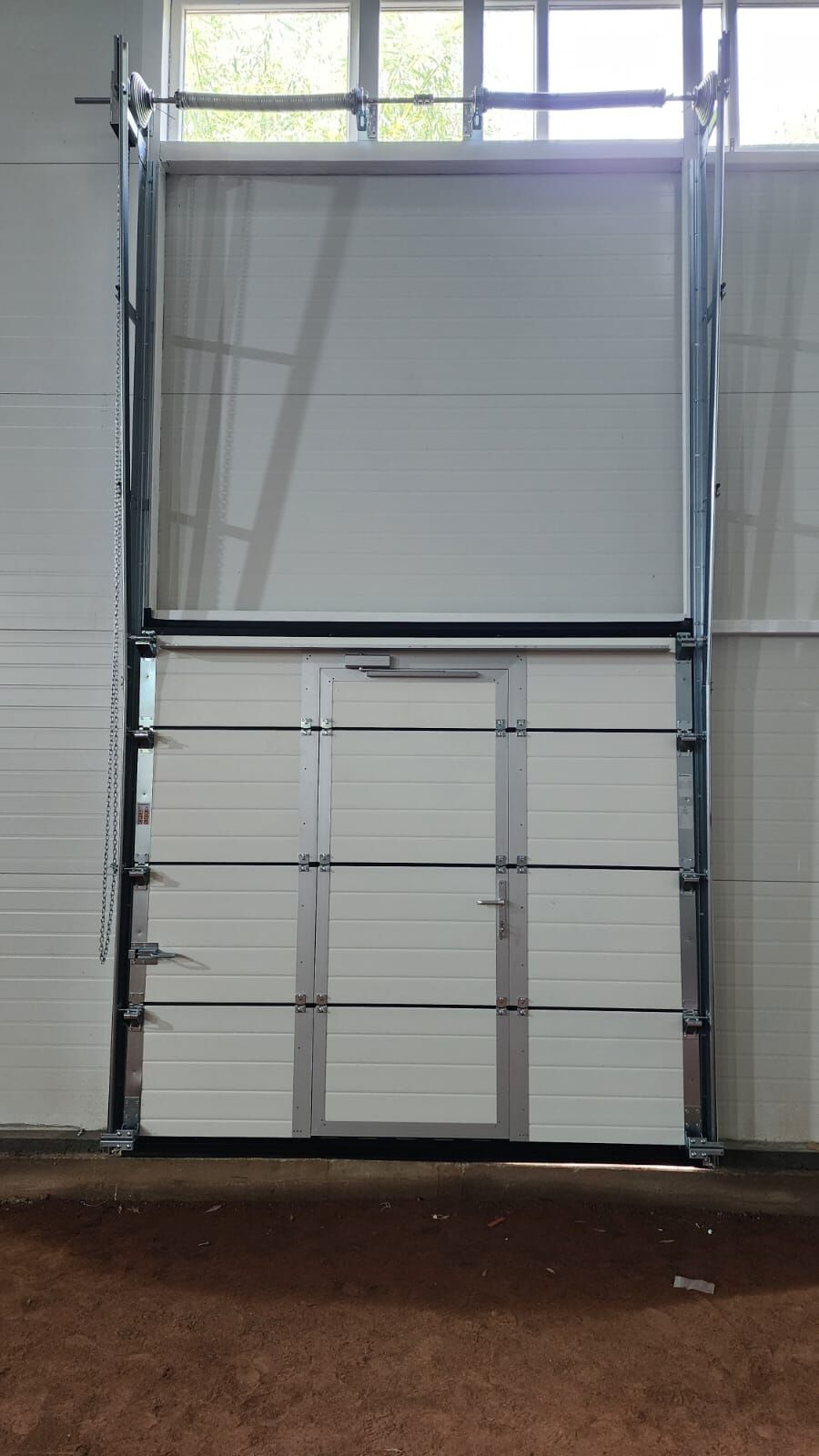 Ușă secțională industrială cu ușă pietonală integrată, văzută din interior, cu arcuri de torsiune și acționare cu lanț