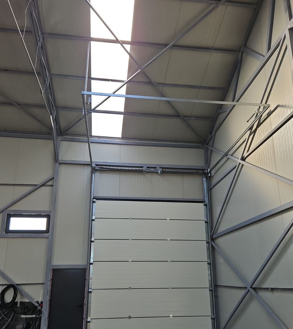 Ușă secțională industrială văzută din interior, cu șine de ridicare înaltă, într-o hală pe structură metalică