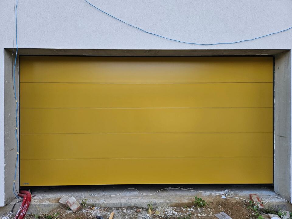 Ușă de garaj secțională galben muștar cu panou neted, montată pe o fațadă albă